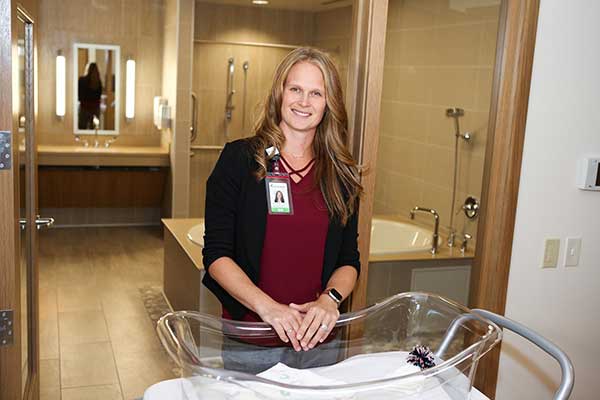 Carrie Lord standing in front of a birthing tub and behind, with her hands resting on, a clear plastic bassinet 