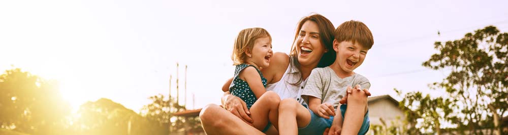 Amother hugging onto her two young children while they all laugh and play outdoors at sunset