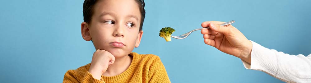 A child looking a parent holding out a piece of broccoli on a fork with a repulsed look on his face
