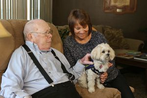 An elderly man and a middle-aged woman sitting on a couch at Tomah Helath Hospice petting a small dog