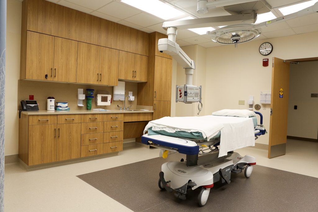 An examining room with cabinets and a sink behind a hospital bed that has a mechanical light hanging above it