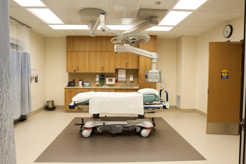 An examining room with cabinets and a sink behind a hospital bed that has a mechanical light hanging above it