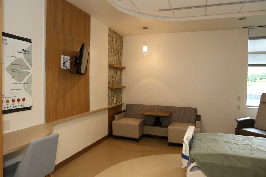 a large patient room at Tomah Health with chairs and a sofa with a built in table and a TV on the wall