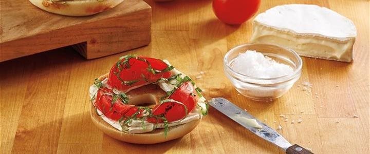 Smoked Tomato, Basil and Brie on a Bagel on a table next to a bowl of salt, butter knife and wedge of brie cheese