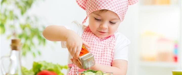 A small child with a chef's hat and apron pourng salt onto food