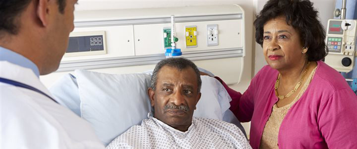 A patient in a hospital gown sitting in a bed with his wife standing next to him talking to the doctor