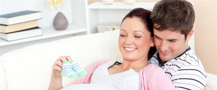 A pregnant woman sitting on and being embraced by a man is holding small baby shoes