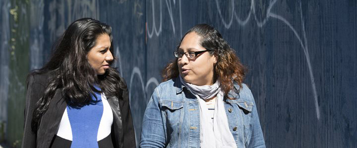 Two women talking to each other while walking past a concrete wall with graffiti