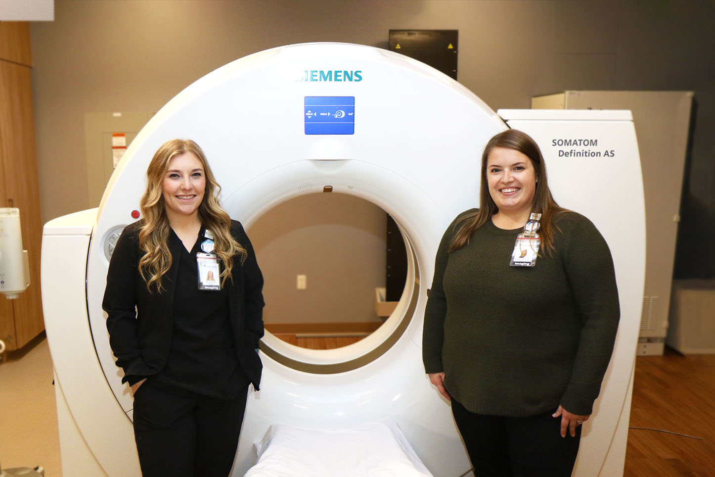 Olivia Aleckson, Imaging Services Technologist, and Alyssa Bronk, Imaging Services Coordinator, pose in front of a CT scanner