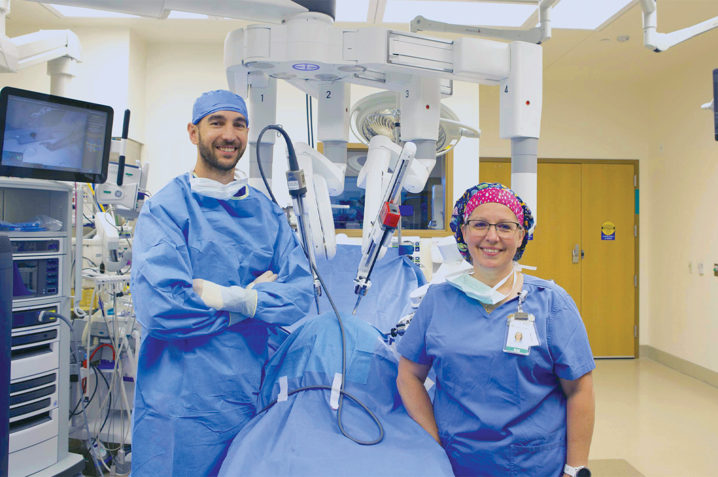 A surgeon and surgery director standing in an operating room on either side of a gurney with a surgical robot behind them