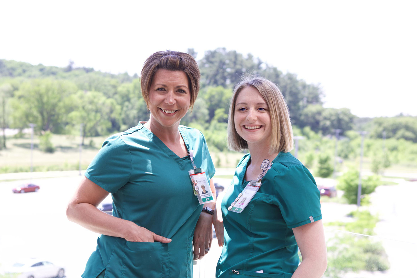 Two nurses standing in front of a large window that overlooks trees in the distance on a sunny summer day