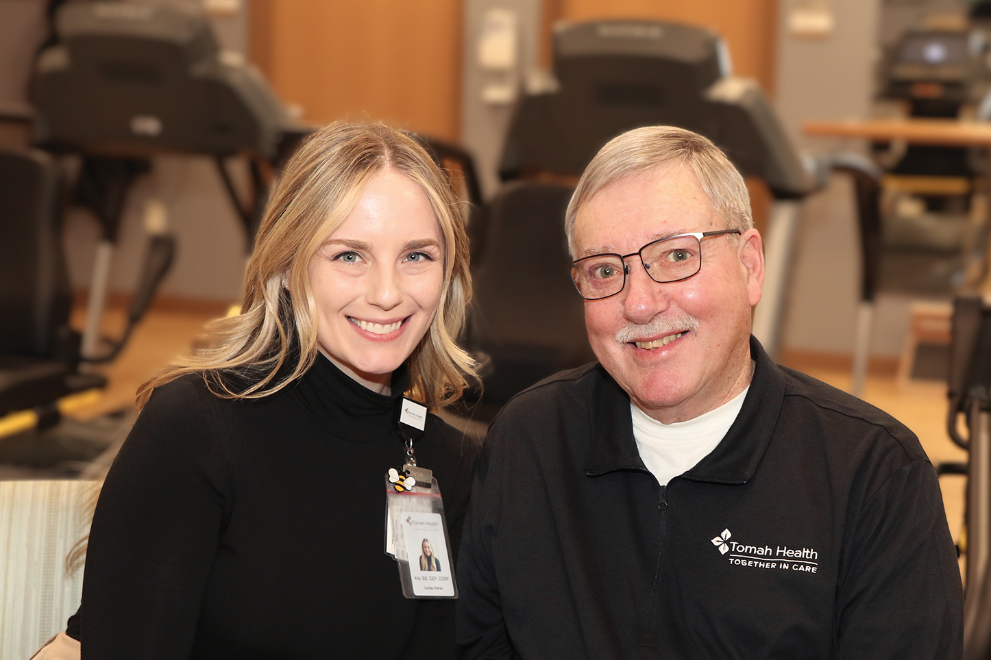Alis Zellmer, Clinical Exercise Physciologist, and Tom Zastoupil, Cardiac Rehab Patient, smile at the camera in the cardiac rehab exercise room at Tomah Health.